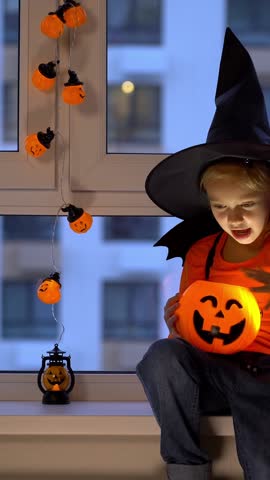 Happy child sits by window in witch hat and conjures over halloween pumpkin lantern in evening. Vertical shot
