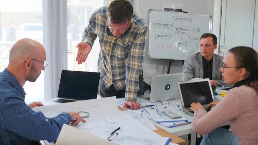 Bald project architect points with a ruler at construction drawings, highlighting nuances to a builder during a heated office discussion. Professional debate over architectural plans.