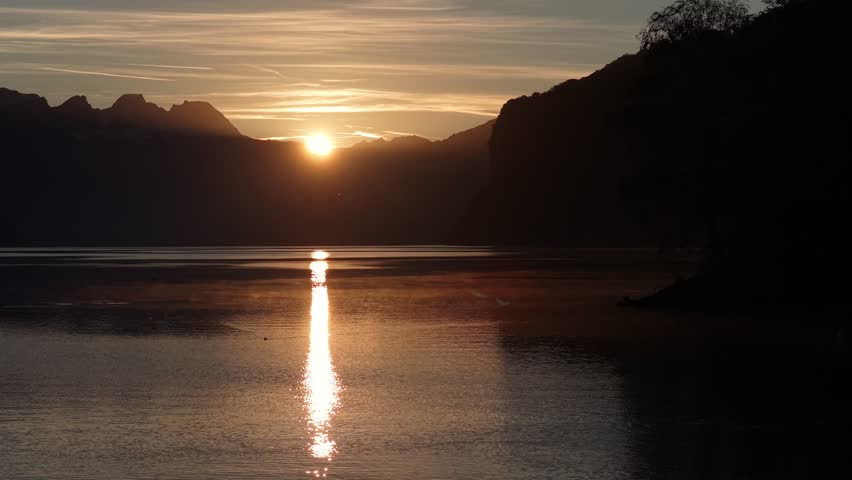 Golden sunset reflecting on a calm lake surrounded by mountains, creating a peaceful and cinematic atmosphere.