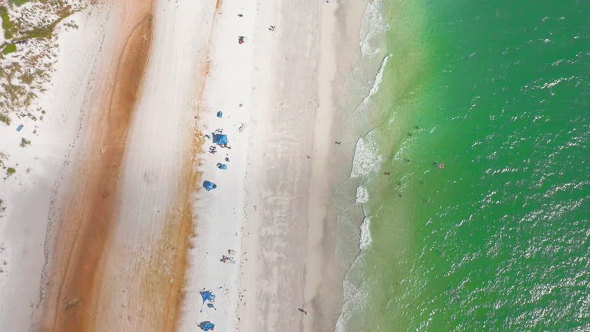 From above, Anna Maria Island stretches along turquoise Gulf waters, its white sandy beach lined with colorful umbrellas, swimmers, and homes nestled between palm-lined streets and the sea.