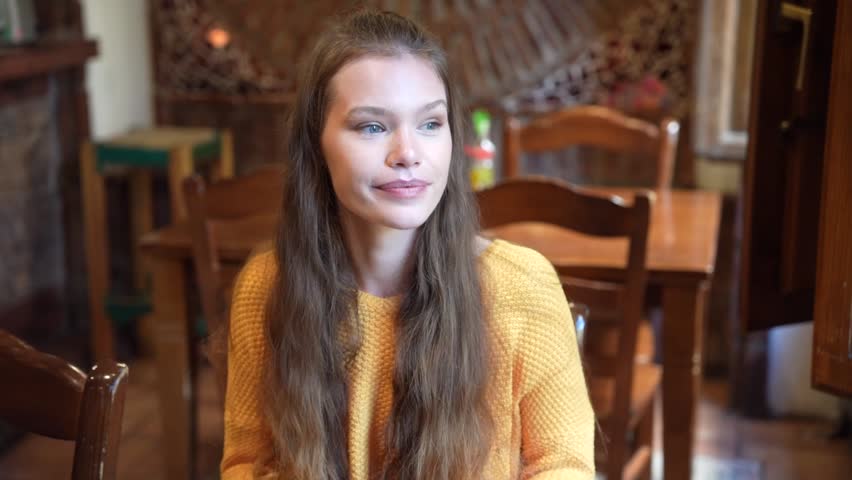 A young woman with long hair sits in a charming caf, wearing a bright yellow sweater, and smiling softly. The warm atmosphere enhances her inviting presence