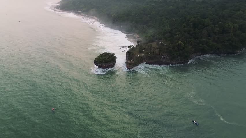 Drone shot of a tropical coastline featuring a lush green forest meeting the ocean. Waves crash against the shore and small rocky islands. Two boats are seen in the water.