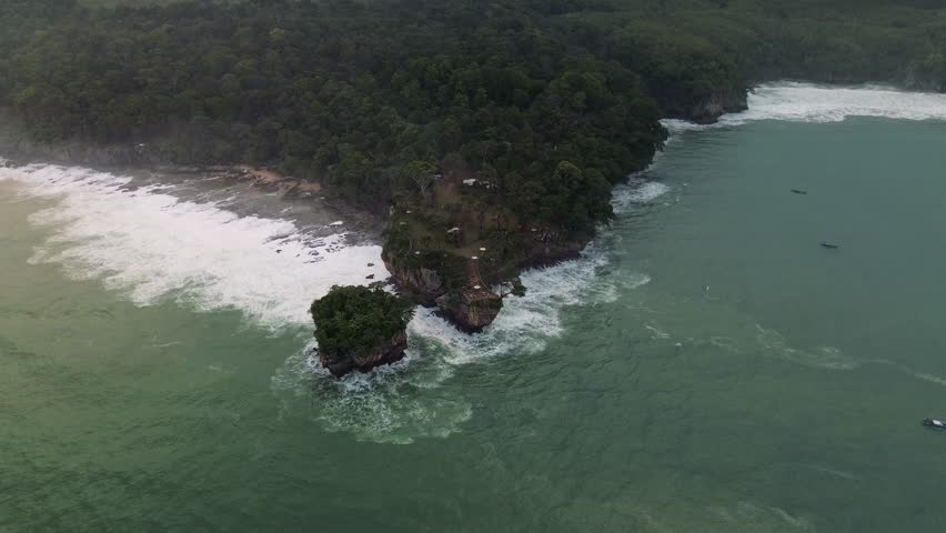 Drone footage captures a lush tropical island with waves crashing against rocky cliffs. A dense forest covers the island, and small boats dot the turquoise sea.