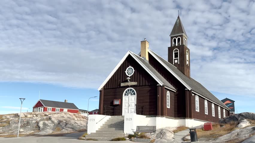 A wooden church in Greenland showcases its historic architecture. The building features a tall spire and beautiful windows, set in a natural landscape.