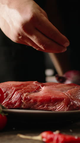 Vertical slow motion video chef hand seasoning raw beef steak with sea salt on kitchen table black background gourmet cooking concept fine dining premium meat restaurant preparation cinematic light