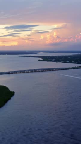A long bridge stretches across a tranquil river as the sun sets behind distant clouds, casting warm reflections of gold, pink, and violet light over the peaceful water below.