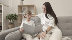 Seven year old boy reads a book while his mother listens to him, sitting together on a gray sofa in a bright cozy living room, warm family and home atmosphere - Powered by Shutterstock - Get 15% off with code: PIKWIZARD15