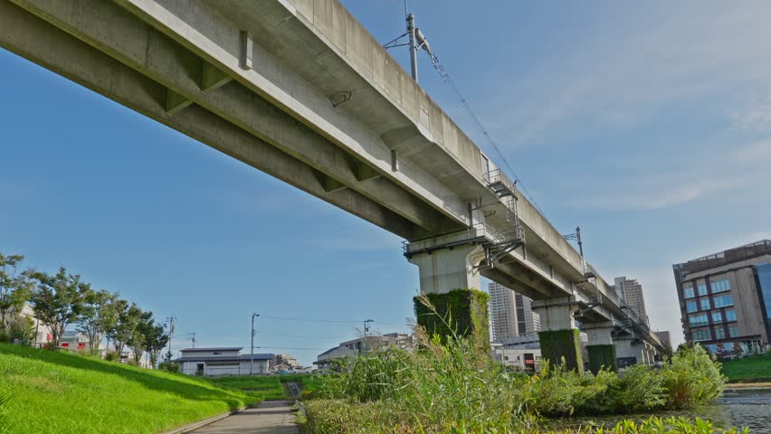 The concrete rail structure of the Tsukuba Express line against a bright blue sky, with modern buildings nearby.