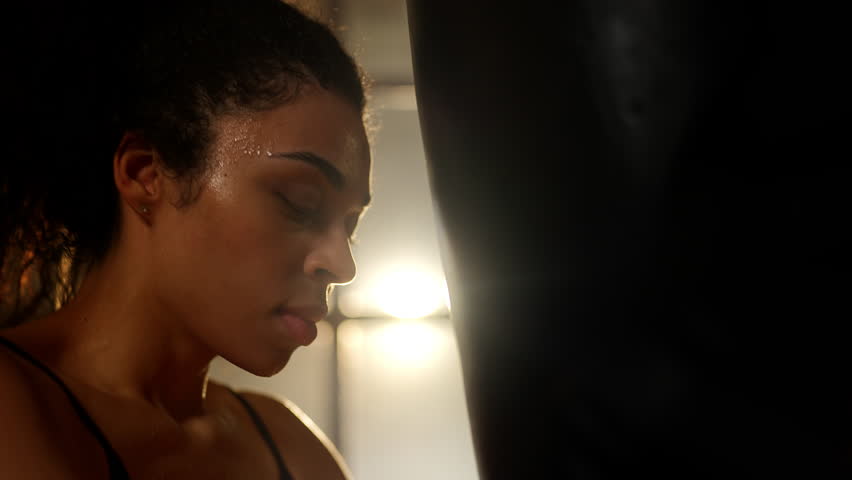 Tired Black female boxer leaning on punching bag after active gym training