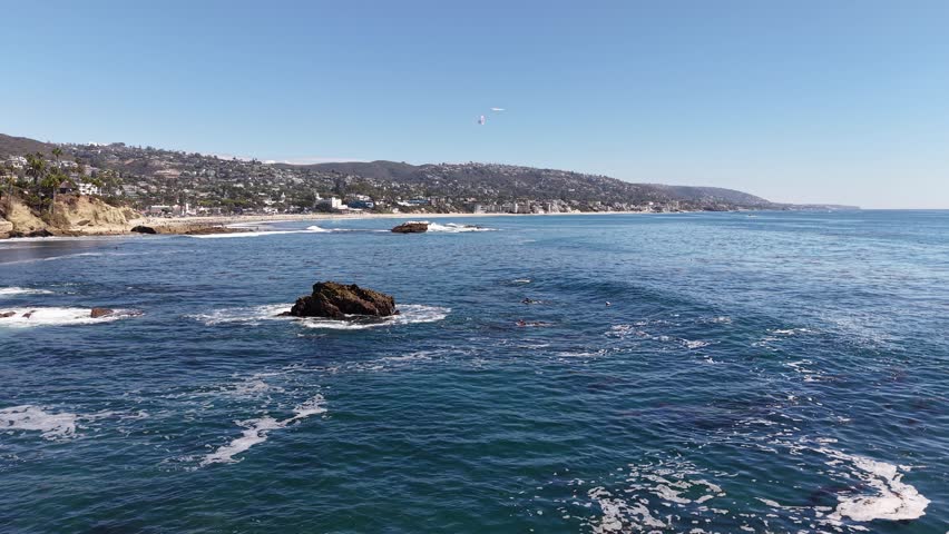Drone captures open ocean and waves meeting the coastline near a rocky outcrop off Laguna Beach, California, under clear skies