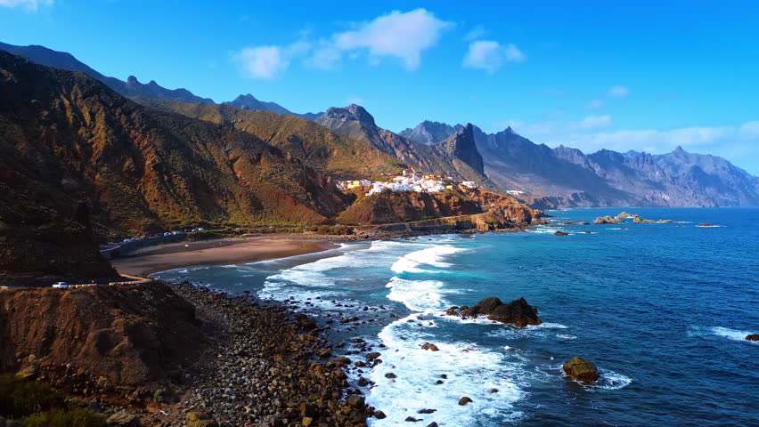 Beautiful foamy waves roll to the narrow sandy beach surrounded by stunning rocks. View on the group of white villas on the rocky shore at backdrop. Tenerife, the Canary Islands, Spain.