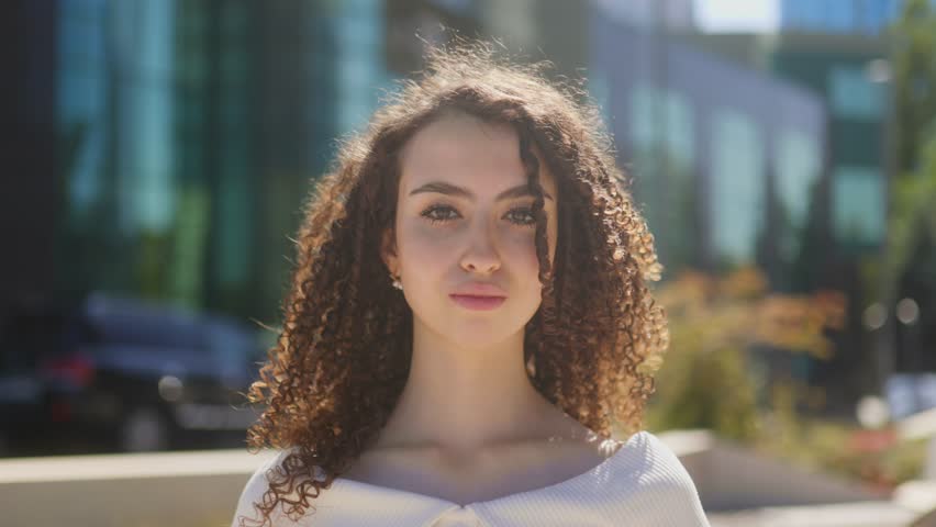 Portrait of a young woman with curly brunette hair looking at the camera on a sunny urban street. Wearing a white blouse, she radiates confidence and natural beauty.