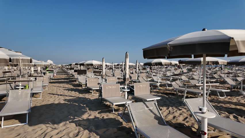 A wooden walkway leads through rows of empty beach chairs and closed parasols on a deserted sandy beach in Rimini, Italy, under a blue sky. High quality 4k footage