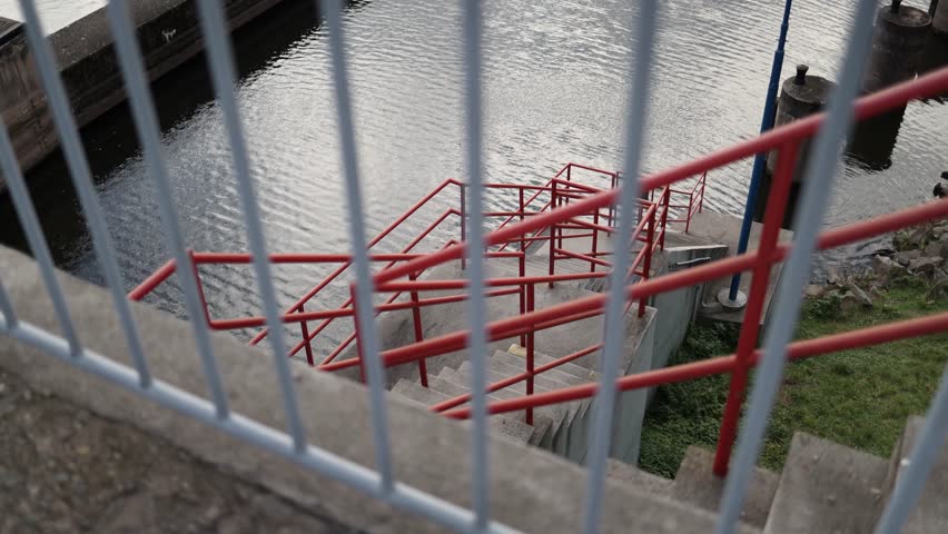 Red metal stairway descends to river through geometric railings viewed from above industrial construction detail with reflections on water and textured concrete creating urban symmetry