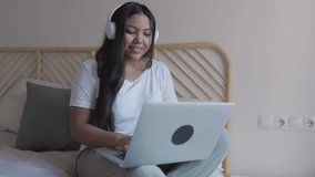 Young asian woman wearing headphones sitting on a bed while using her laptop to work from home - Powered by Shutterstock - Get 15% off with code: PIKWIZARD15