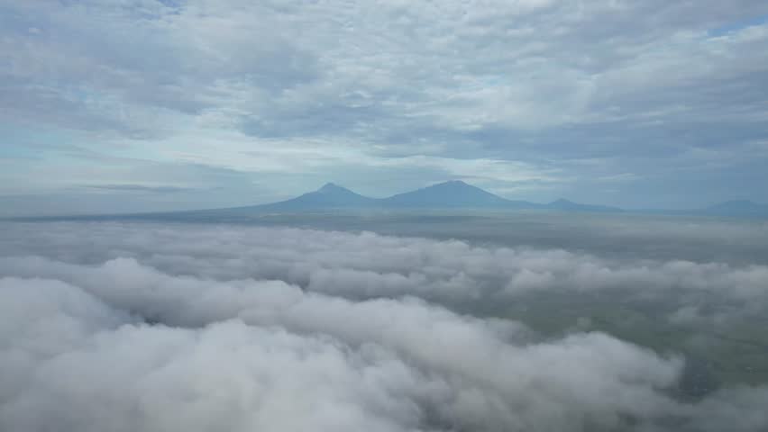 “Drone flies straight toward Mount Merbabu and Mount Merapi in Boyolali, Central Java, Indonesia, capturing the blue-tinted mountains under a clear sky.”