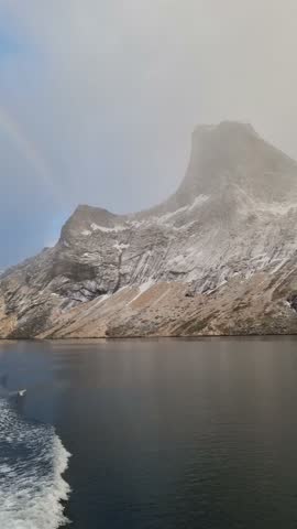 Beautiful snowy mountains over fishing village Reine in Lofoten islands, Norway, with partially frozen fjord and rainbow in winter at sunrise. Dramatic sky over snowy rocks and sea at dawn
