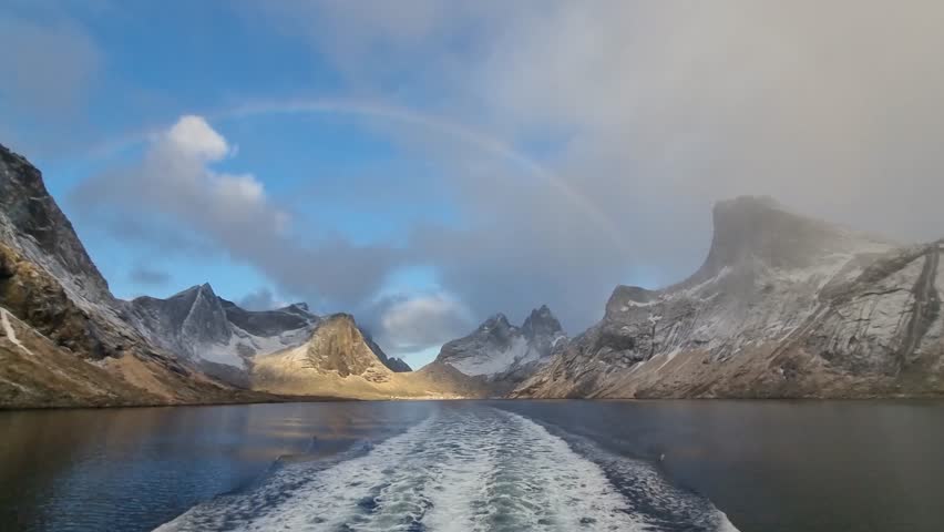 Beautiful snowy mountains over fishing village Reine in Lofoten islands, Norway, with partially frozen fjord and rainbow in winter at sunrise. Dramatic sky over snowy rocks and sea at dawn
