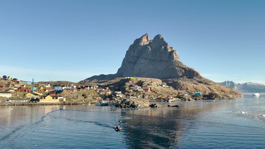Brightly colored houses sit along a peaceful fjord in Greenland. Snowy mountains rise in the background, creating a stunning natural landscape with icebergs floating in the water.