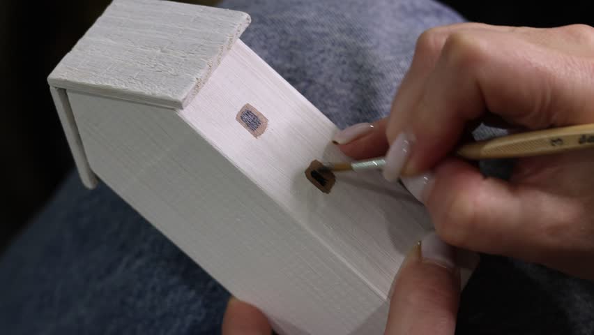 Captivating close-up of a woman expertly painting a miniature wooden house window frame with a fine brush, adding intricate details to her handcrafted creation indoors, displaying precision.