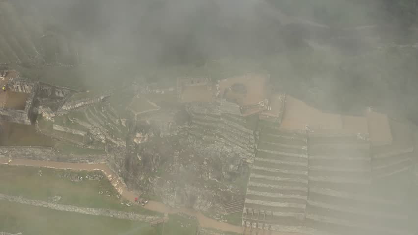 Aerial shot moves at a low altitude across Machu Picchu, offering a close, downward-facing view of the intricate stone walls, structures, and multi-level terraces as heavy mist and clouds drift