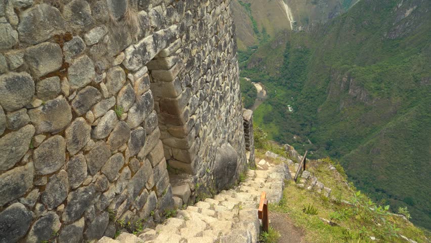A dramatic shot looking straight down from the peak of Huayna Picchu, showing the narrow, steep stone stairs carved into the mountainside that lead up to the final upper citadel and sacred structures 