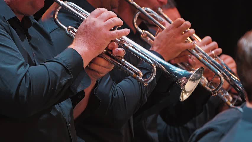 Close-up of musicians playing trumpets in a brass ensemble. The focus is on the hands gripping the instruments, showcasing the shiny metal and the musicians dressed in black attire