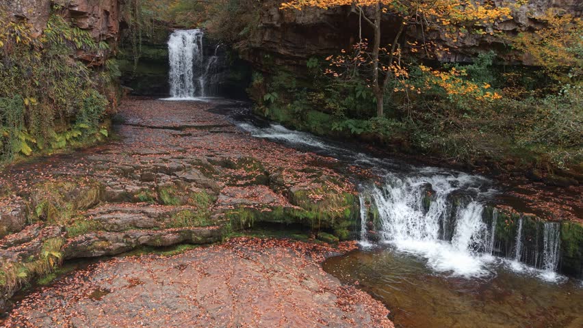 Drone flying towards a beautiful waterfall in a Welsh woodland during autumn