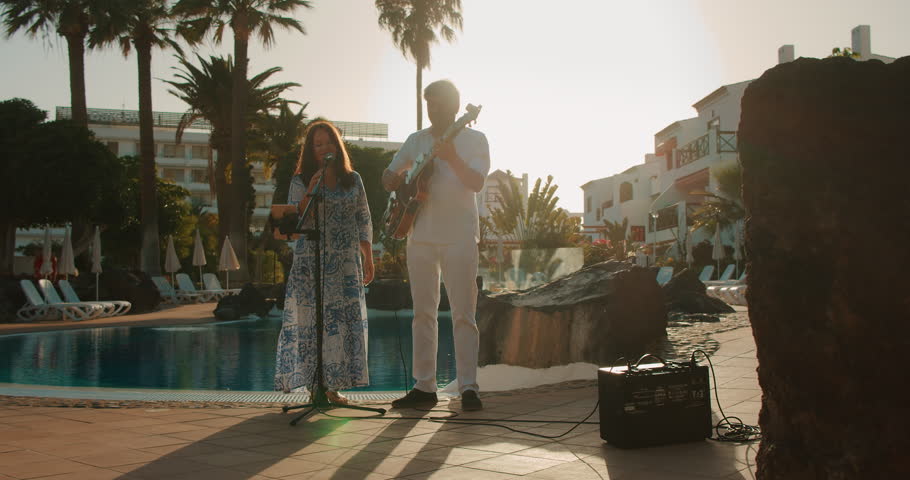 Two musicians perform poolside at sunset with guitar and microphone setup
