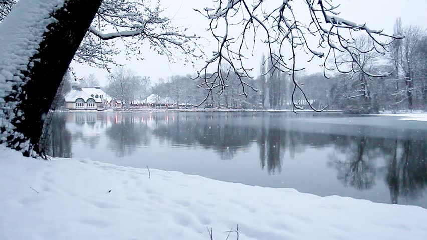 Beautiful fluffy snow falling over a dark lake in a city park during winter. Everything around is covered with white snow, creating a calm and magical winter atmosphere. English Garden park in Munich
