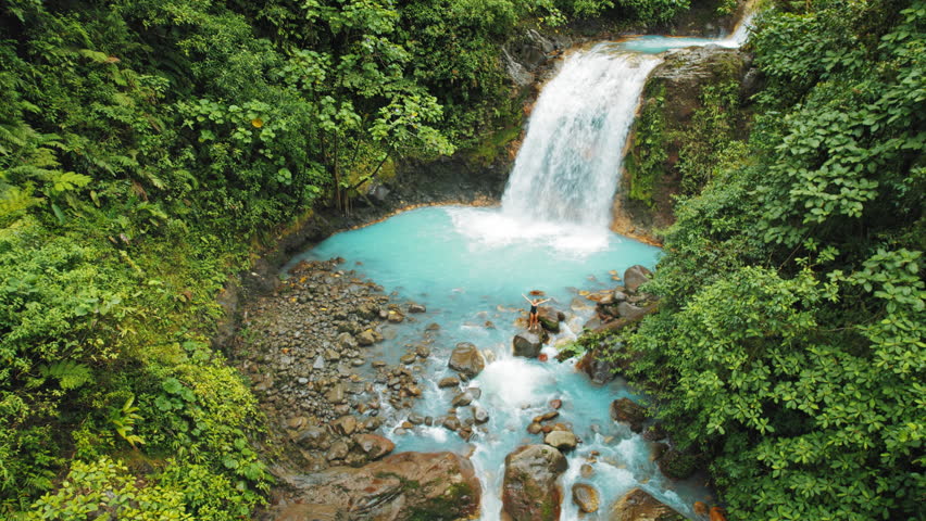 Woman standing on rock near turquoise pool below waterfall in Costa Rica jungle