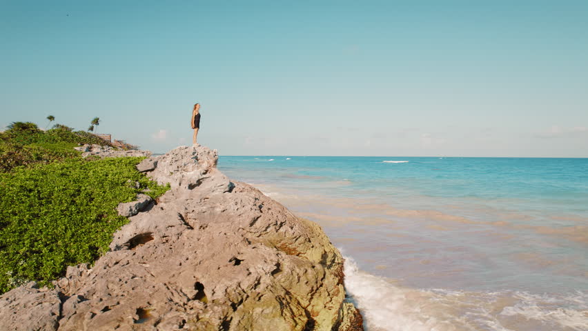 Woman standing on rocky cliff by the ocean looking out to sea in Tulum, Mexico