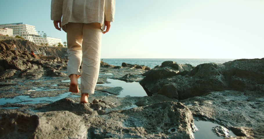 Person walking barefoot on volcanic rocks near ocean at sunset