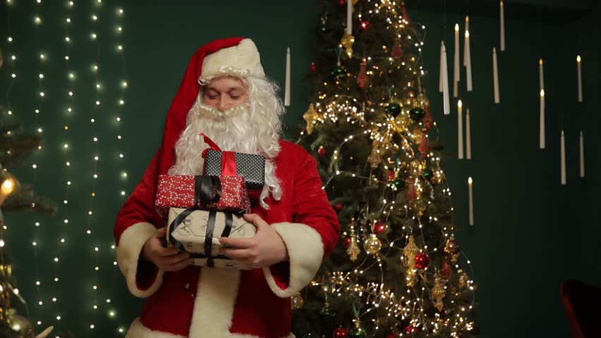 Santa holding wrapped gifts near a Christmas tree with festive lights.