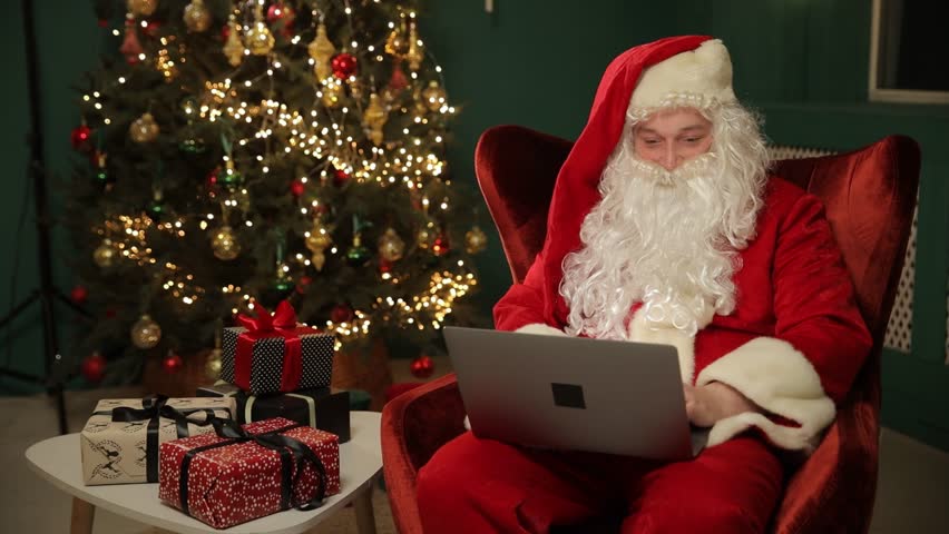 Santa in cozy studio reviewing gift list on laptop near Christmas tree.