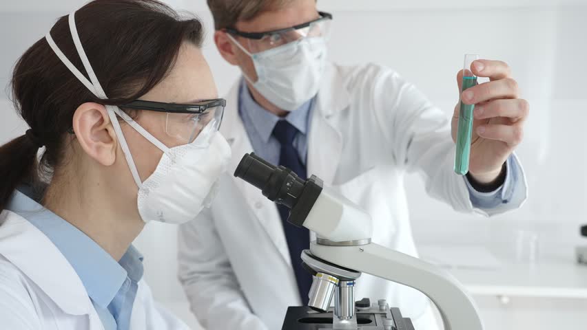Two scientists in lab coats and safety gear conducting research, one observing through a microscope and the other holding a test tube with turquoise liquid. Medicine and science concept