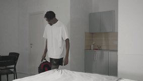 Man in white t-shirt bending down to pick up backpack in minimalist bedroom, with green headboard, bed with neatly folded towels, and clean modern decor in the background - Powered by Shutterstock - Get 15% off with code: PIKWIZARD15