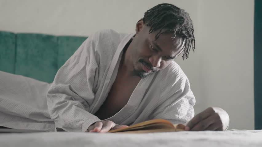 Man in white robe lying on bed, deeply focused on reading large book, hand turning pages slowly. Soft light from window creates peaceful, contemplative atmosphere in calm indoor setting