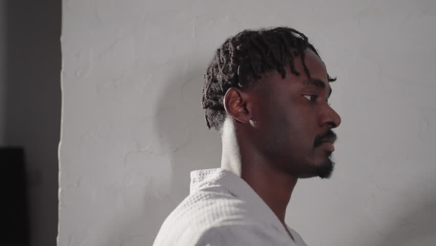 Close-up of man in bathrobe holding spoon and preparing to stir a drink in clear glass cup. Modern kettle and minimalist kitchen design visible in background, soft morning light enhancing the scene