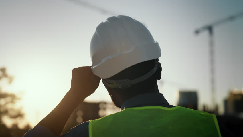 Builder walking at construction site, back view against sunset sky, slow motion. Middle-age male person correcting protective helmet on head, foreman or architect examining process of modern building