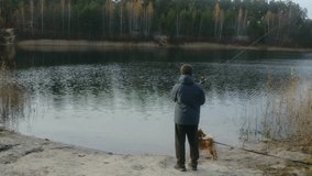 Young man wearing a jacket and hat fishing with a spinning rod from the sandy shore of a scenic lake, with his loyal shiba inu dog standing patiently beside him on a leash - Powered by Shutterstock - Get 15% off with code: PIKWIZARD15