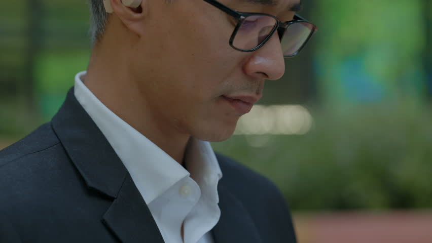 Portrait of mid aged Asian businessman with hearing aid types on laptop as sits outside office closeup. Chinese entrepreneur with disability using computer in downtown. Accessibility at workplace