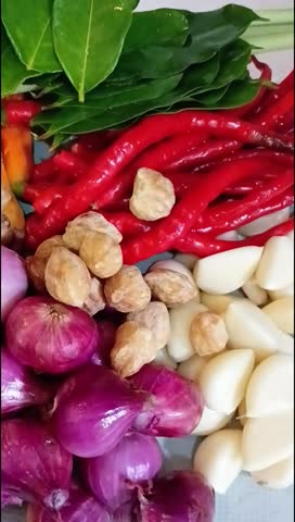 Close-up displays raw red peppers, garlic cloves, red onions, green leaves and seed pods prepared for cooking or food photography usage.