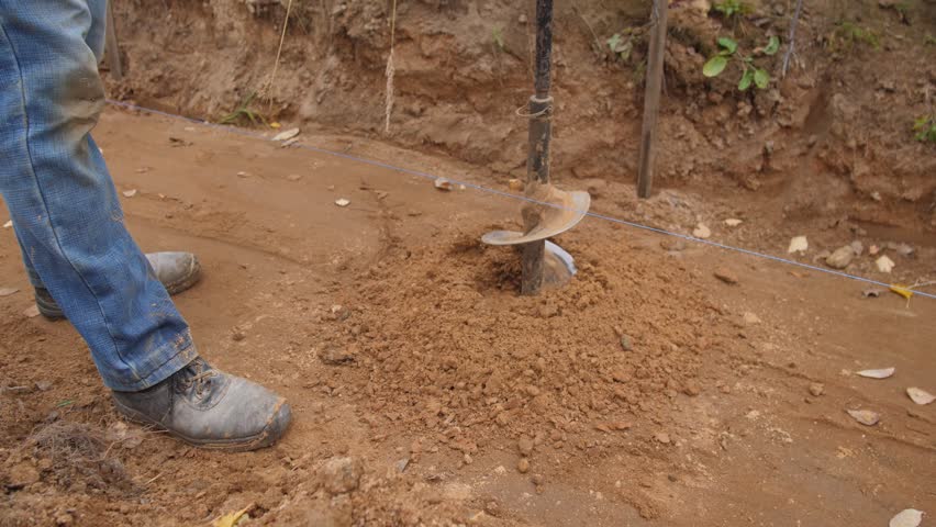 man smoothing earth with tools, laborer carefully levels loose soil during rural drilling operation, worker using boots and auger to smooth and compact soil around drilled hole in rural
