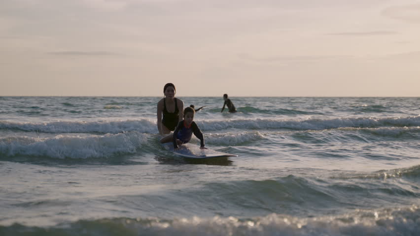 Happy little boy learning to play with a surfboard from his mother, having fun and passion in the sea during summer vacation at the beach. An Asian mom teaches and trains her cute son on how to surf. 