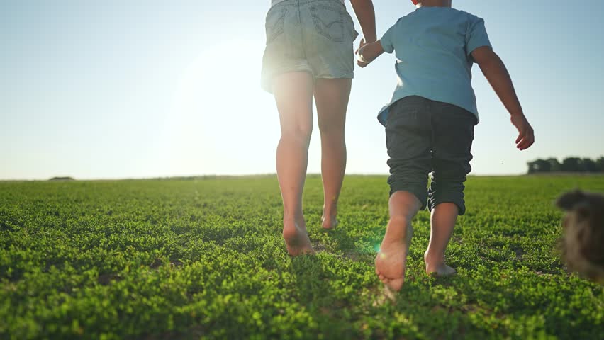 Barefoot children walking together on green grass in summer field. Girl and boy holding hands while walking. Dog runs happily near children in grassy field. Summer barefoot walk with holding hands.