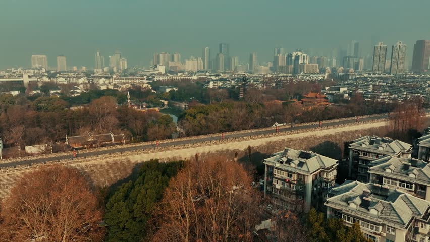 Nanjing, China. Aerial view of historic cityscape with stone wall and river, and construction workers on elevated walkway. The scene is set against a backdrop of a hazy sky.
