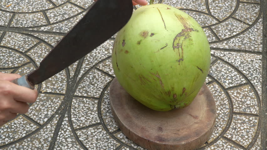 A person's hands use a large knife (machete) to skillfully chop open a fresh green coconut on a wooden block. Outdoor, traditional method of preparing a tropical drink.