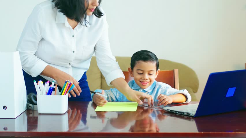 A woman is helping a young boy with his homework. The boy is sitting at a desk with a laptop and a stack of papers. The woman is holding a pair of scissors and a pen, and she is pointing to the papers