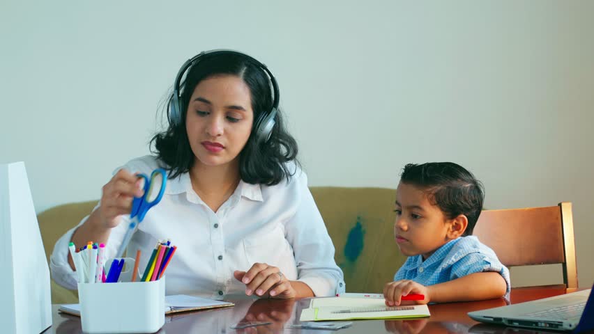 A woman is helping a child with a school assignment. The child is sitting at a table with a laptop and a notebook. The woman is holding a pair of scissors and a pen